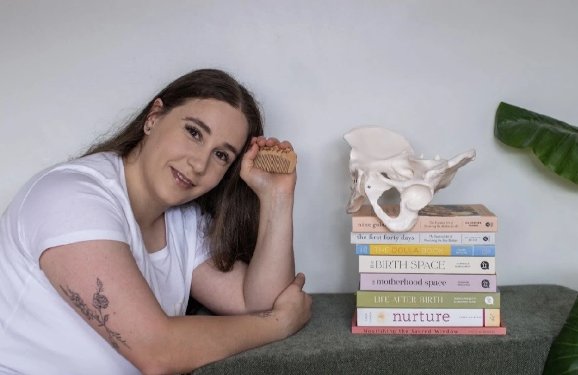 Doula Helene of Gaia Nourishment sitting with a comb in hand next to birth books with a bone pelvis on top